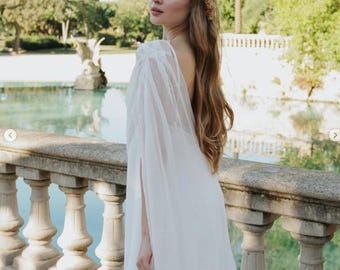 Puede incluir: Un vestido blanco largo hasta el suelo con mangas transparentes tipo capa y espalda descubierta, combinado con una delicada corona de flores con pequeños pétalos rosas y naranjas, lucido por una persona de cabello largo y ondulado.