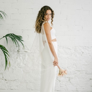 May include: A woman wearing a white bridal top and trousers with a sheer white cape. She is holding a small bouquet of dried flowers.