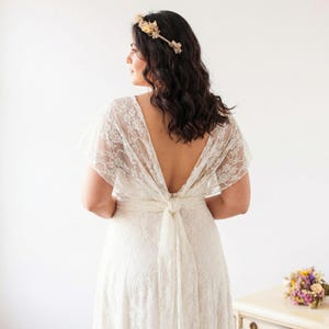 May include: A white lace dress featuring a deep V-back design with a tied sash at the waist, short flutter sleeves, and a floral lace pattern throughout. The garment is paired with a delicate floral crown made of small, light-colored blossoms.