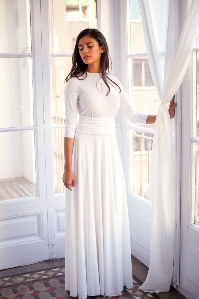 The model is wearing a long wedding dress with three-quarter sleeves while touching the white curtains on her left.