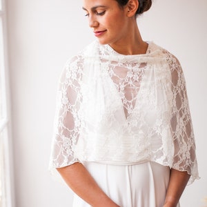 May include: A white lace bolero shrug featuring a delicate floral pattern and short sleeves, worn over a simple white sleeveless dress. The garment is lightweight and semi-sheer, with a high neckline and a scalloped edge along the hemline.