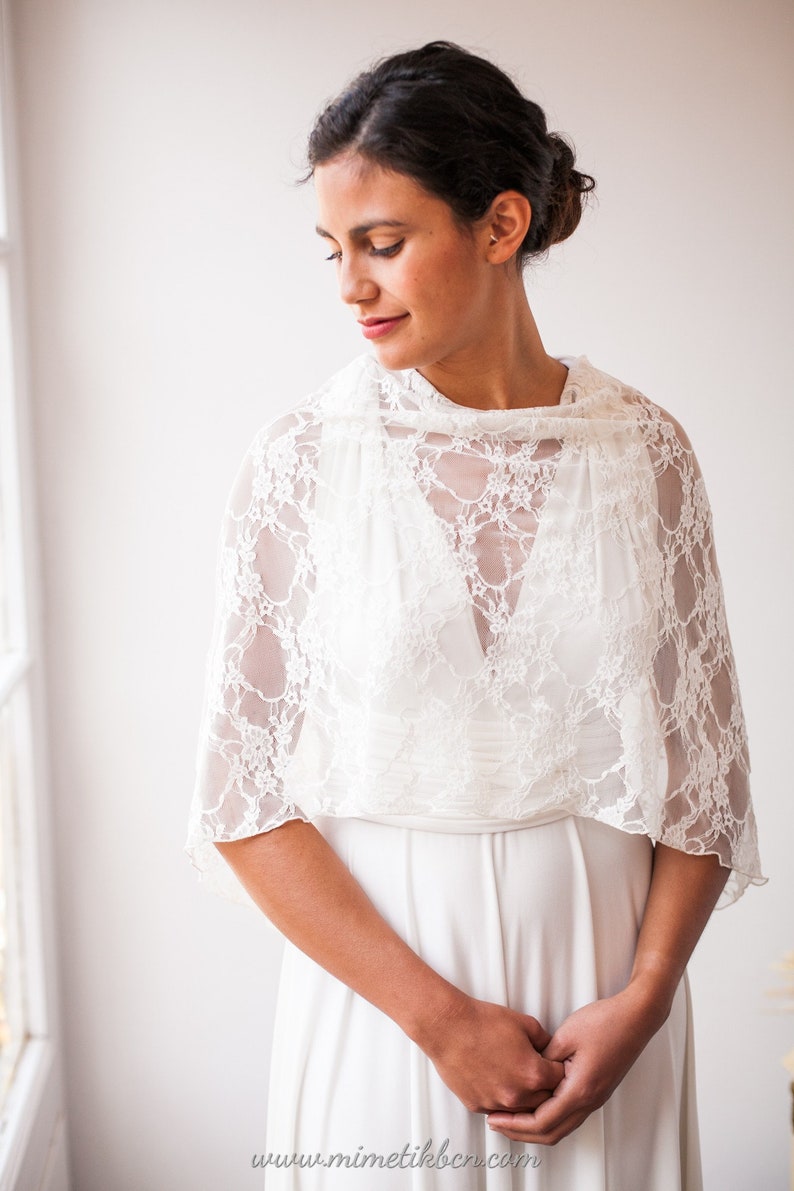 May include: A white lace bolero shrug featuring a delicate floral pattern and short sleeves, worn over a simple white sleeveless dress. The garment is lightweight and semi-sheer, with a high neckline and a scalloped edge along the hemline.