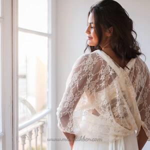 May include: An ivory lace bolero with three-quarter length sleeves and a delicate floral pattern. The garment features a draped back design with a crossover detail, worn over a plain white dress. The text www.mimetikbcn.com is visible.