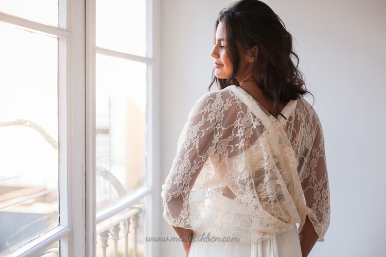 May include: An ivory lace bolero with three-quarter length sleeves and a delicate floral pattern. The garment features a draped back design with a crossover detail, worn over a plain white dress. The text www.mimetikbcn.com is visible.