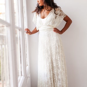 A woman wears a wedding dress in a modernist flat, with big windows. The model is next to the window. She has her head slightly turned. The woman is wearing a grecian wedding dress. The lace of the dress is off-white.