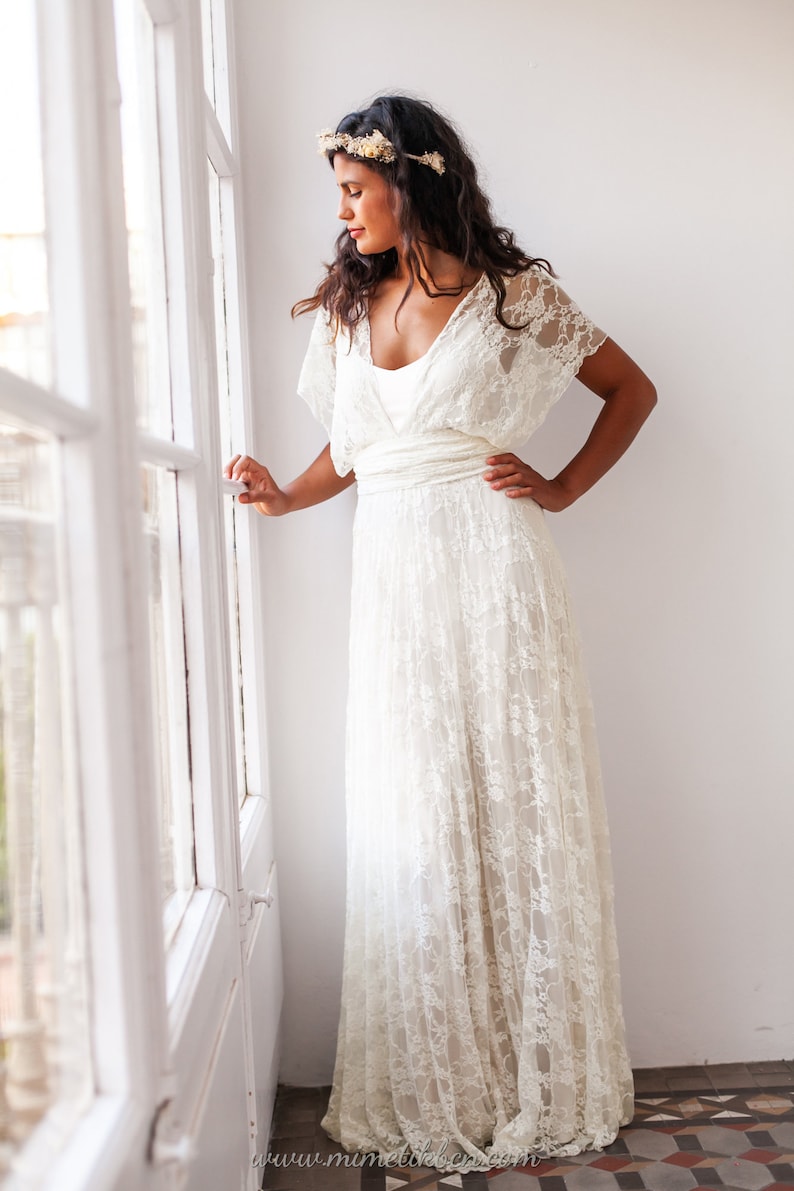 A woman wears a wedding dress in a modernist flat, with big windows. The model is next to the window. She has her head slightly turned. The woman is wearing a grecian wedding dress. The lace of the dress is off-white.