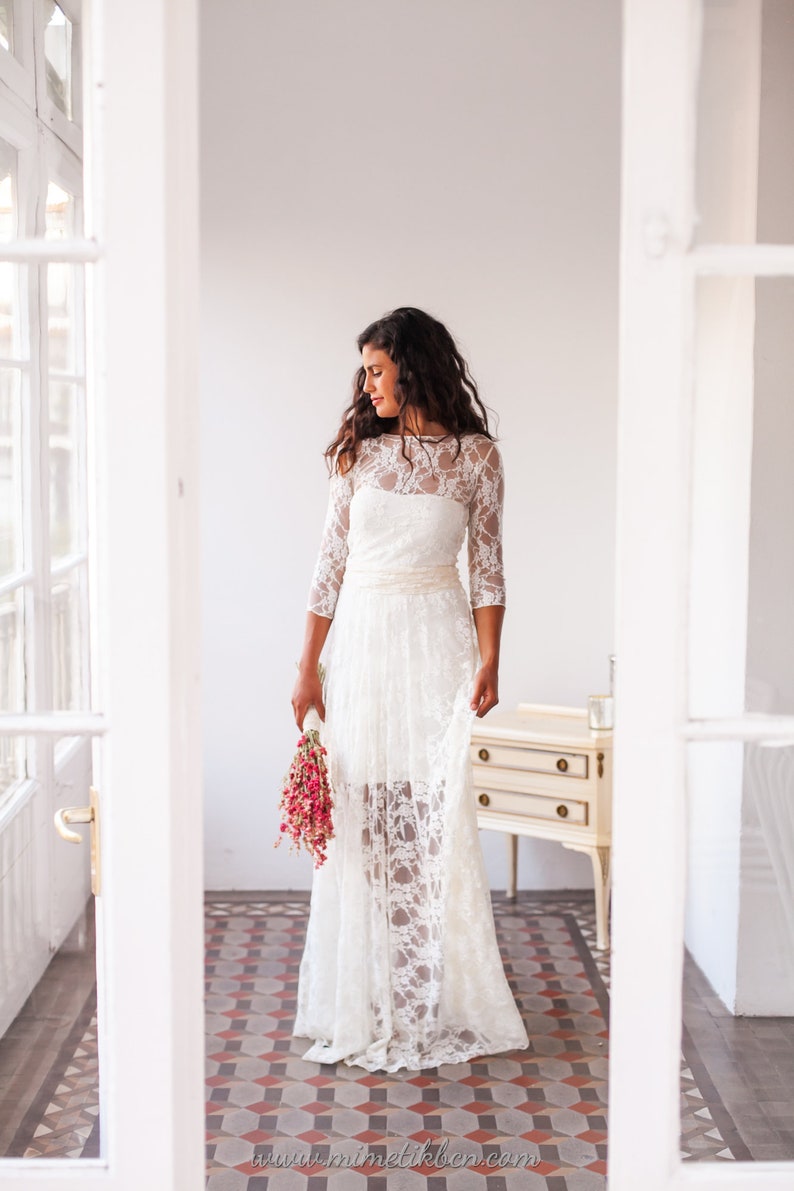 May include: A floor-length white lace dress featuring three-quarter length sleeves, a round neckline, and a solid white bodice underlay. The skirt has a sheer lace overlay with a scalloped hemline and a wide, textured waistband in a cream hue.