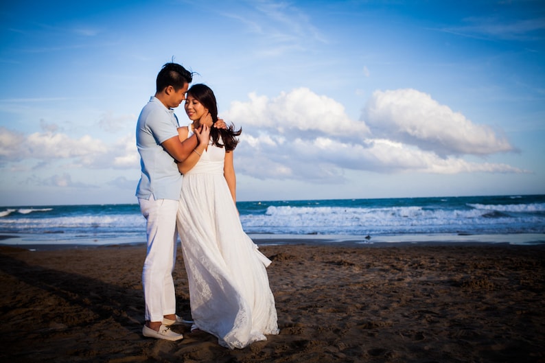 May include: A person wearing a light blue polo shirt and white trousers stands next to a person wearing a long white sleeveless dress with a lace hem. They are positioned on a sandy beach with the ocean and a blue sky with clouds in the background.