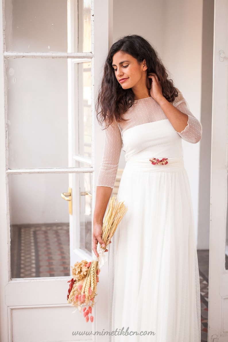 May include: A white floor-length dress featuring a sheer dotted mesh overlay on the bodice and three-quarter length sleeves, a solid white waistband, and a floral accent at the waist. The text www.mimetikbcn.com is printed at the bottom.