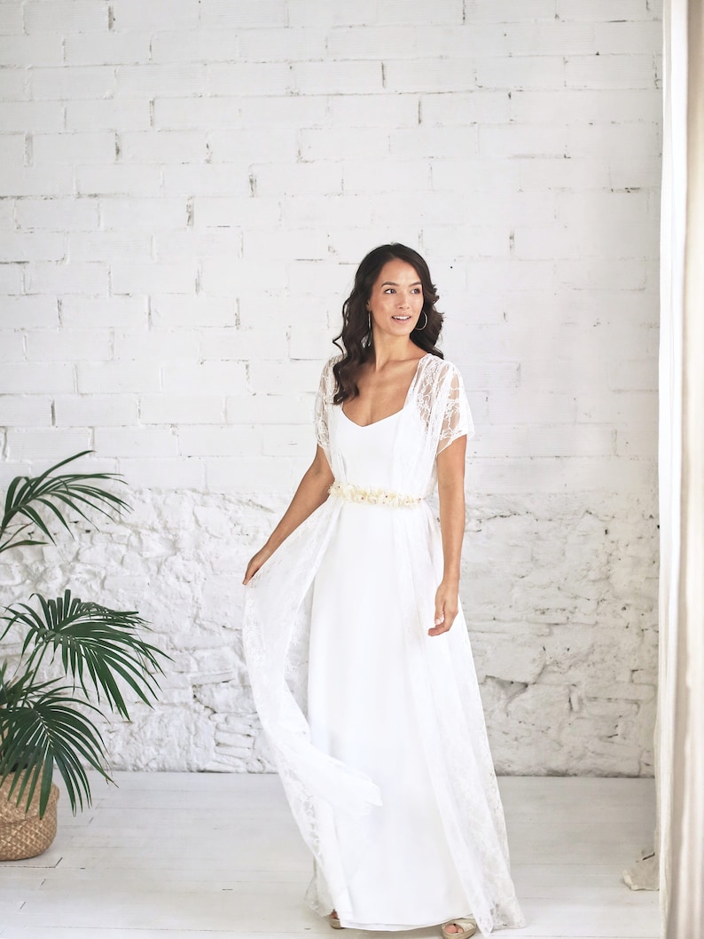 May include: A white lace wedding dress with a flowing skirt and a floral belt. The dress has a simple, elegant design and is perfect for a beach wedding.