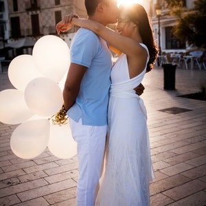 May include: A light blue polo shirt, white trousers, and beige loafers paired with a white floor-length dress featuring a lace overlay. A cluster of white balloons with gold ribbons is held by the individual wearing the light blue polo shirt.