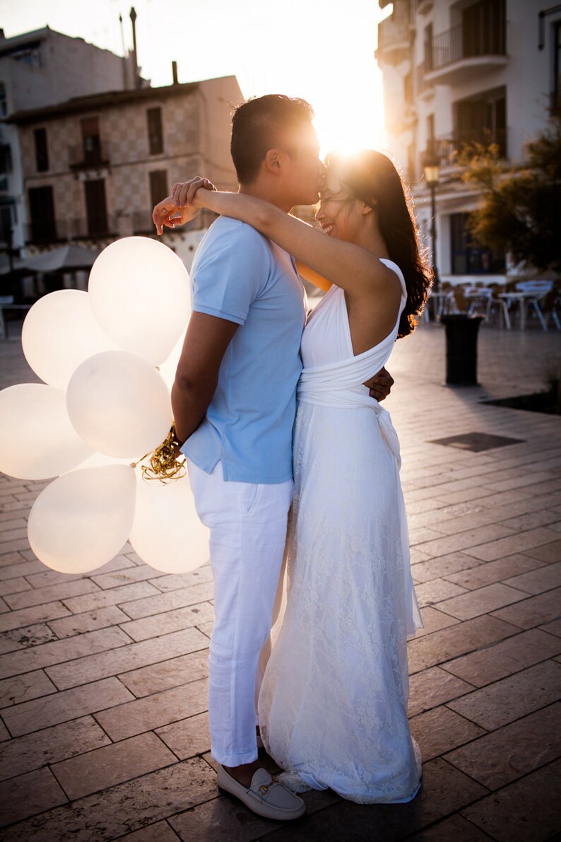 May include: A light blue polo shirt, white trousers, and beige loafers paired with a white floor-length dress featuring a lace overlay. A cluster of white balloons with gold ribbons is held by the individual wearing the light blue polo shirt.