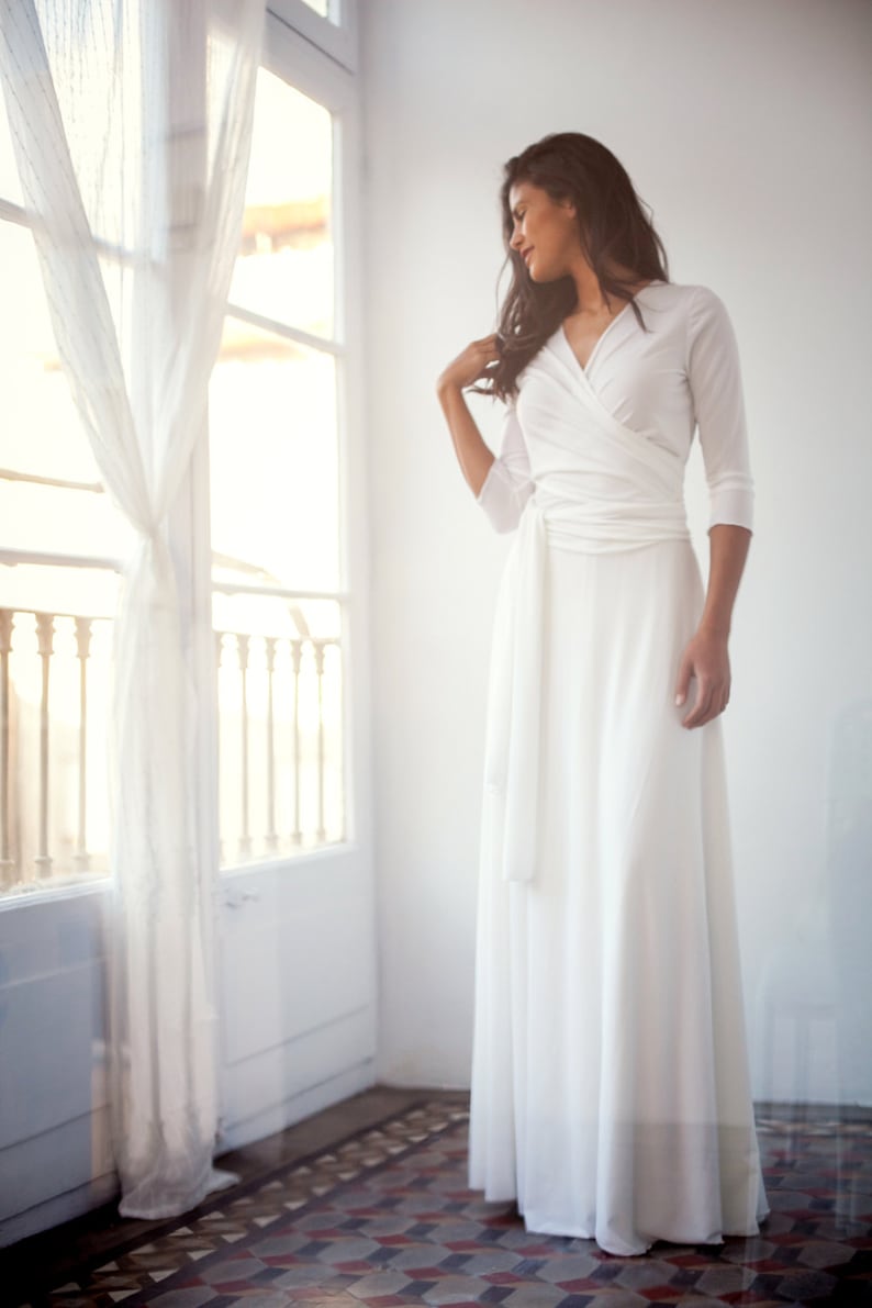 Woman wearing a simple white wrap dress as a wedding dress. The bridal gown is long and has a deep v-neck. She is staring outside while smiling.