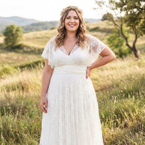 May include: A floor-length white lace dress featuring a V-neckline, short flutter sleeves, and a fitted waistband. The dress is adorned with an intricate floral lace overlay throughout the bodice and skirt, paired with a gold-toned floral crown.