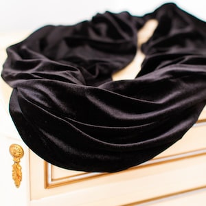 May include: A black velvet infinity scarf with a soft, light-reflecting texture and deep, draped folds. The fabric has a smooth, plush surface that creates subtle highlights and shadows across the circular, continuous loop design.