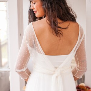 May include: A white dress featuring three-quarter length sleeves made of sheer dotted swiss fabric. The garment has a deep scoop back neckline with a fabric tie closure at the waist. The text www.mimetikbcn.com is printed at the bottom.