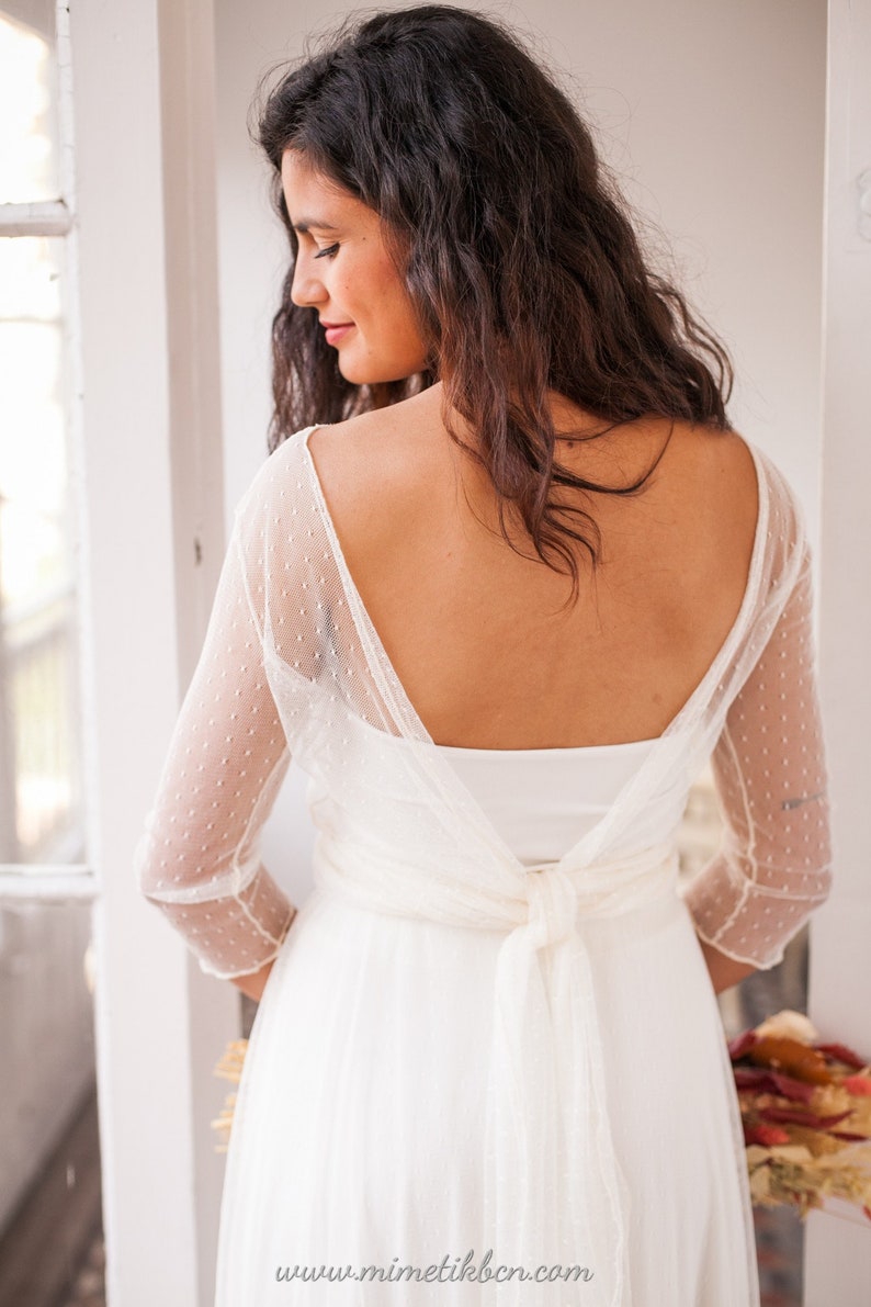 May include: A white dress featuring three-quarter length sleeves made of sheer dotted swiss fabric. The garment has a deep scoop back neckline with a fabric tie closure at the waist. The text www.mimetikbcn.com is printed at the bottom.