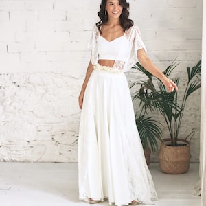 May include: A white lace bridal top with a sweetheart neckline and a matching white lace skirt. The skirt is long and flowing, and the top is cropped. The model is wearing a flower crown.