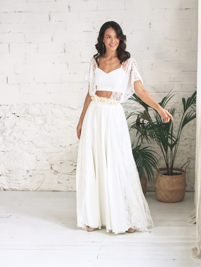May include: A white lace bridal top with a sweetheart neckline and a matching white lace skirt. The skirt is long and flowing, and the top is cropped. The model is wearing a flower crown.