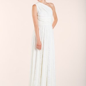 May include: A floor-length white dress featuring a one-shoulder bodice with wrapped fabric detailing at the waist and a long, flowing skirt with a subtle textured pattern. The garment is sleeveless and constructed from a solid white fabric.