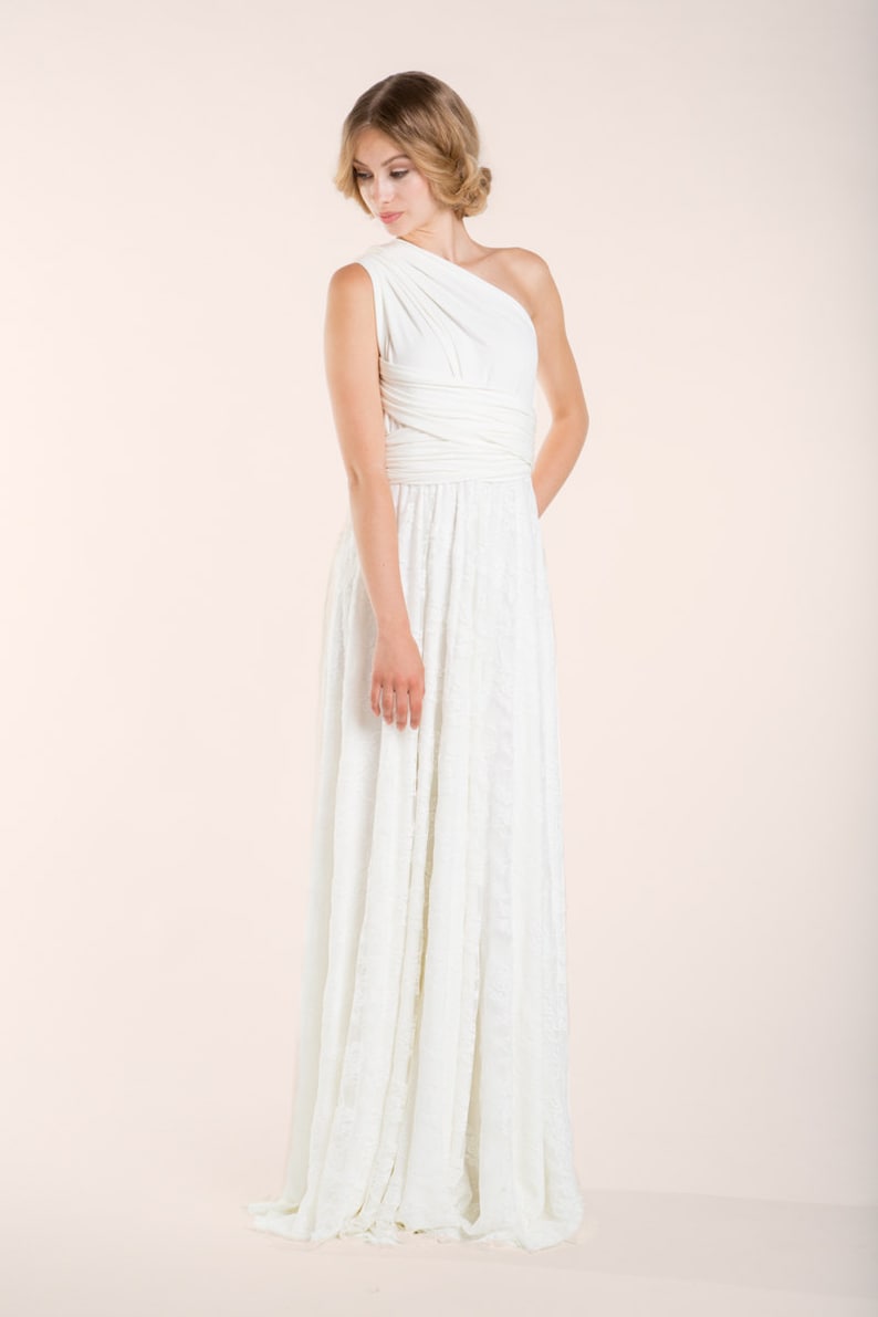 May include: A floor-length white dress featuring a one-shoulder bodice with wrapped fabric detailing at the waist and a long, flowing skirt with a subtle textured pattern. The garment is sleeveless and constructed from a solid white fabric.