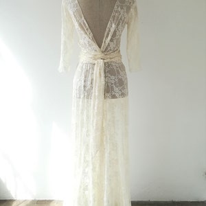 May include: A floor-length cream-colored lace dress featuring three-quarter length sleeves, a deep V-neckline, and a wide fabric tie at the waist. The sheer floral lace pattern covers the entire garment, which is displayed on a dress form.
