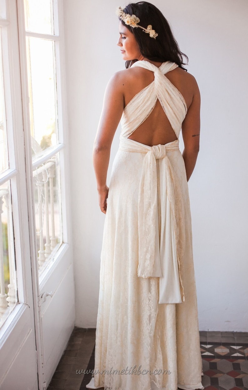 May include: A floor-length cream-coloured dress featuring a lace overlay, a crisscross halter neckline, and an open back design with a tied sash at the waist. A floral headpiece with small white flowers is worn on the head, completing the ensemble.