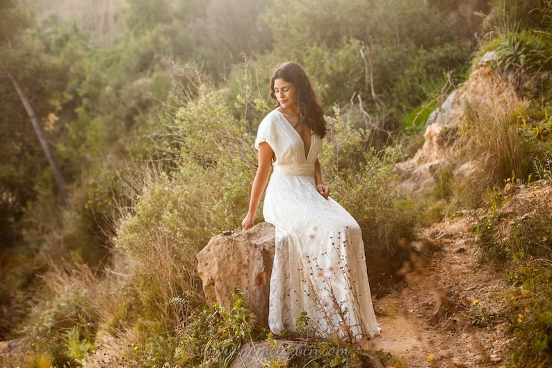 There is a woman in the nature wearing a beautiful wedding dress. The ambiance is relaxed and she looks calm. The dress is made in ivory and ivory lace, creating an elegant complementary look. The dress has floor length, so we cannot see her shoes.