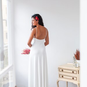 A woman wearing a simple wedding dress. She is giving her back so we can see her discovered and elegant back. The dress has thin straps and is made in an ivory satin jersey.