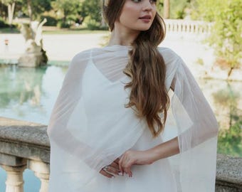 Puede incluir: Un vestido blanco sin mangas con una capa de tul transparente y drapeada que se extiende en mangas fluidas tipo capa. La prenda se combina con una delicada corona floral compuesta por pequeñas flores secas de color rosa y amarillo en la cabeza.