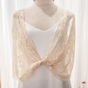 May include: A cream-colored lace bolero with a floral pattern and a knotted front detail, worn over a plain white dress. A matching cream-colored fabric headband with a knotted top sits atop the mannequin head, completing the ensemble.