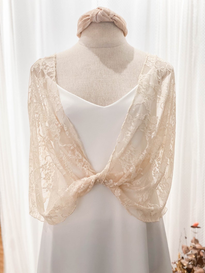 May include: A cream-colored lace bolero with a floral pattern and a knotted front detail, worn over a plain white dress. A matching cream-colored fabric headband with a knotted top sits atop the mannequin head, completing the ensemble.