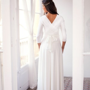 Woman showing her back looking at her left, staring outside through a big window. She has a long white dress with sleeves.