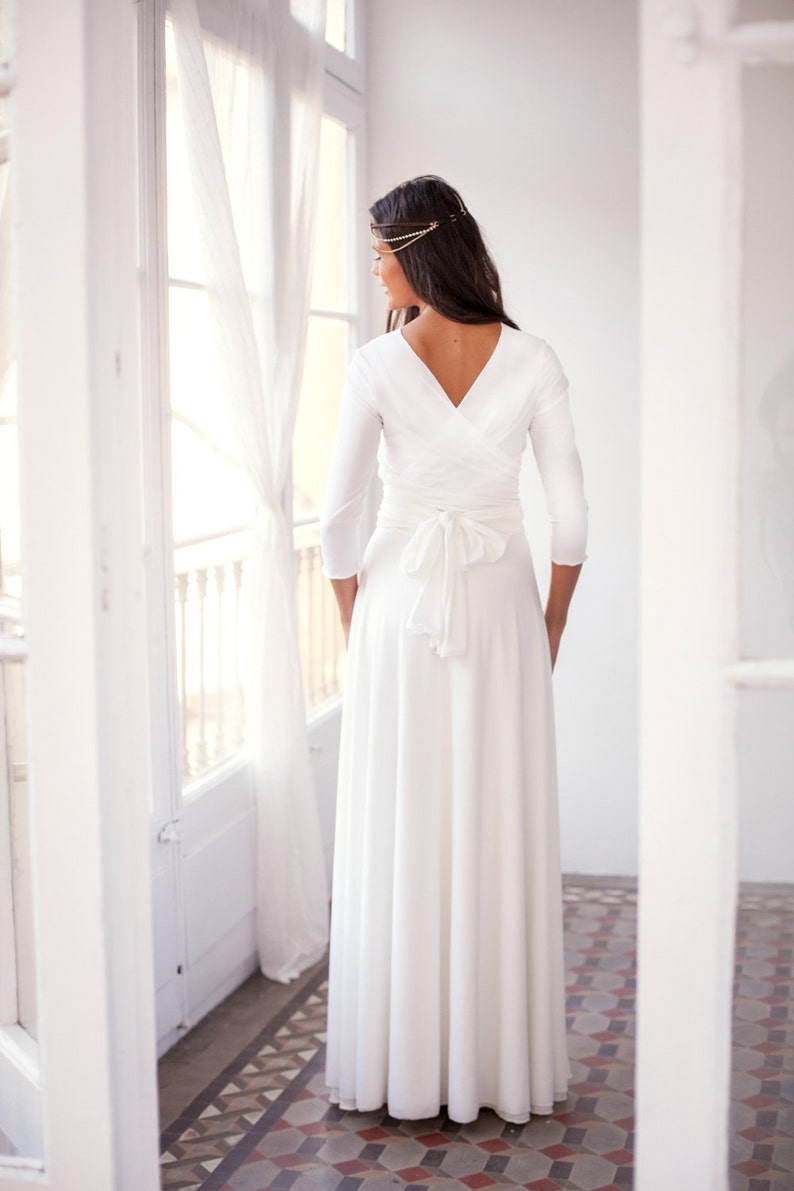 Woman showing her back looking at her left, staring outside through a big window. She has a long white dress with sleeves.