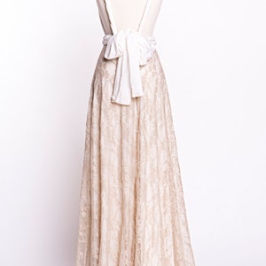 May include: A floor-length dress featuring a beige lace skirt and a solid white bodice. The back of the garment displays thick white straps that cross and secure with a large, prominent white fabric bow at the waistline, creating an open-back design.