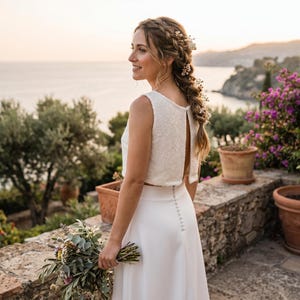 May include: A two-piece white ensemble consisting of a sleeveless crop top with a subtle textured floral pattern and a matching floor-length skirt featuring a vertical row of small white buttons down the center back, paired with a bouquet of wildflowers.