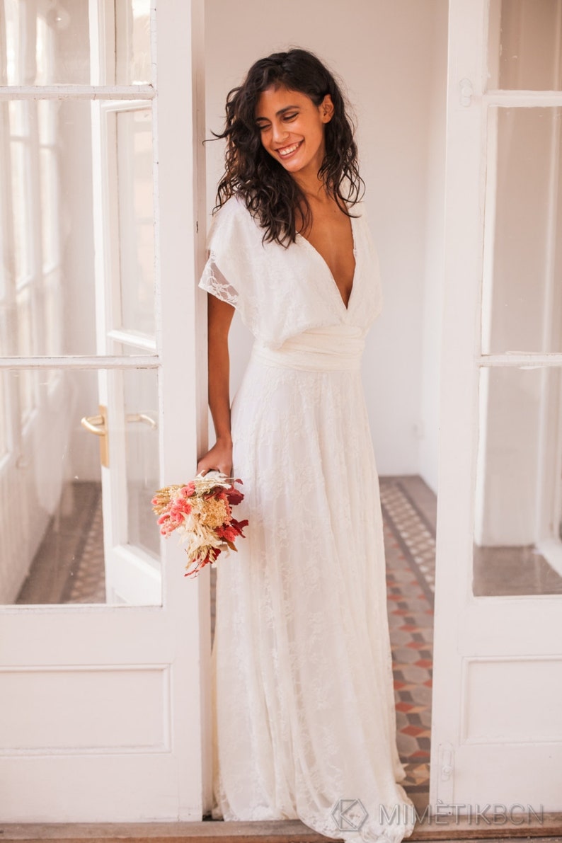 May include: A floor-length white lace dress featuring a deep V-neckline, short flutter sleeves, and a wide fabric waistband. The dress is crafted from a delicate floral lace overlay and is paired with a small bouquet of dried pink and cream flowers.