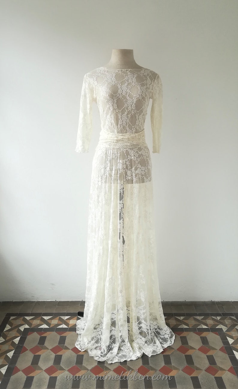 May include: A floor-length, off-white lace dress featuring long sleeves, a boat neckline, and a fitted waist sash. The garment is crafted from a sheer, intricate floral lace pattern that extends from the bodice down to the hemline.