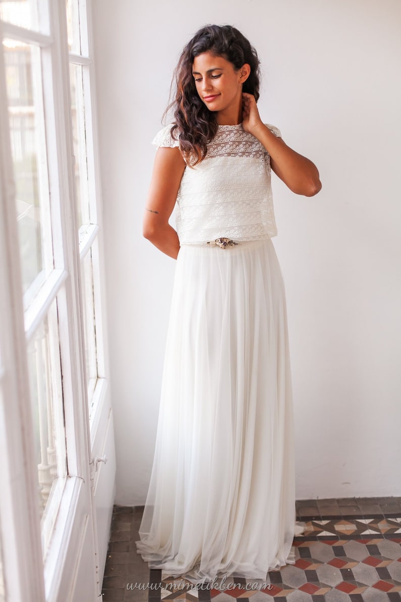 May include: A white lace crop top with a white tulle skirt and a gold belt. The model is standing in front of a white wall with a window behind her.