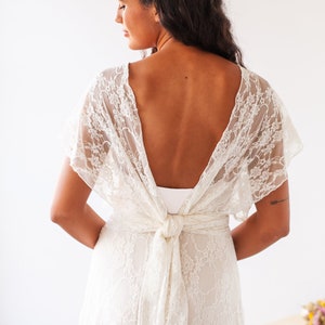 Woman showing the back of her bridal dress. It has short sleeves and is made of ivory lace. Next to her, there's a table with some flowers in it as boho decor.