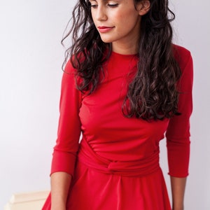 May include: A vibrant red dress featuring three-quarter length sleeves, a round neckline, and a fitted bodice with a gathered, knotted waist detail that transitions into a flared skirt. The fabric appears smooth and solid in color.