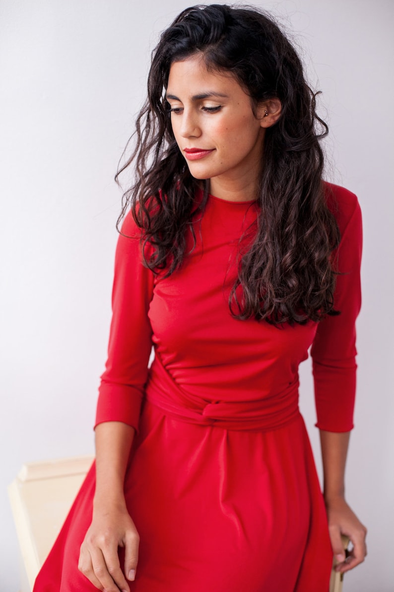 May include: A vibrant red dress featuring three-quarter length sleeves, a round neckline, and a fitted bodice with a gathered, knotted waist detail that transitions into a flared skirt. The fabric appears smooth and solid in color.
