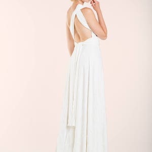 May include: A floor-length white dress featuring a one-shoulder design with a wide strap, an open back, and a long fabric tie at the waist. The skirt flows downward in a straight silhouette, crafted from a solid white fabric with a subtle texture.