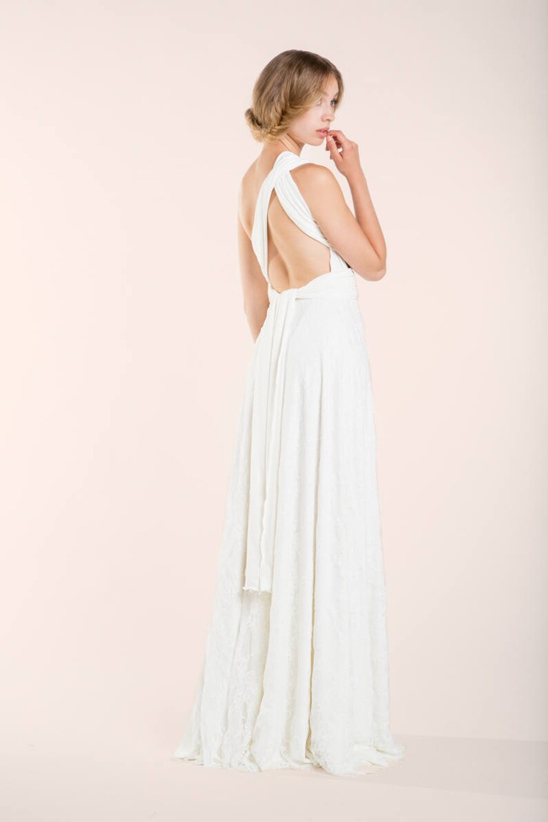 May include: A floor-length white dress featuring a one-shoulder design with a wide strap, an open back, and a long fabric tie at the waist. The skirt flows downward in a straight silhouette, crafted from a solid white fabric with a subtle texture.