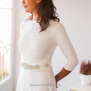 Close-up of the bridal gown. It's eaily to appreciate the vintage belt that combines with the bridal gown. Also, it's easy to see the ivory lace of the overskirt and how elegant the long sleeves are