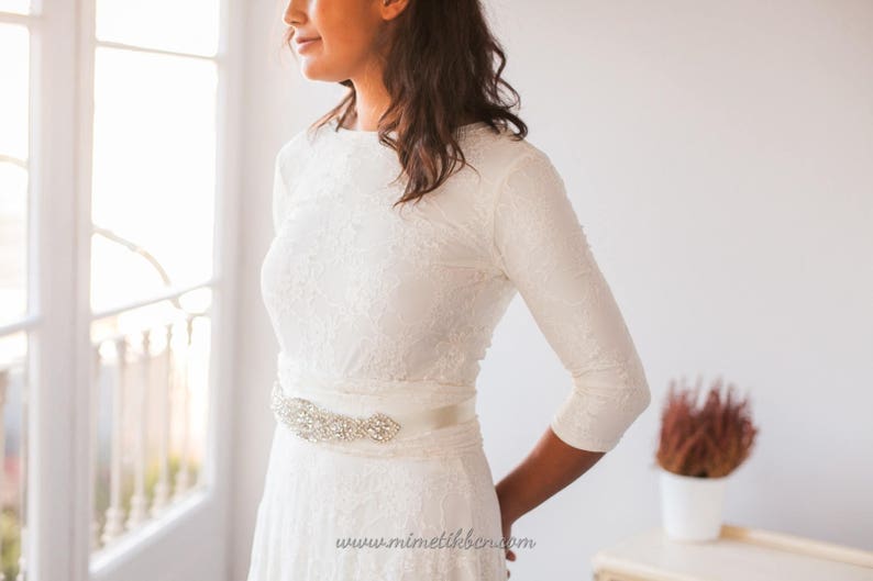 Close-up of the bridal gown. It's eaily to appreciate the vintage belt that combines with the bridal gown. Also, it's easy to see the ivory lace of the overskirt and how elegant the long sleeves are