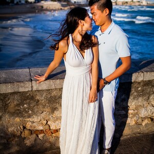 May include: A floor-length white dress with a halter neckline and a lace skirt paired with a light blue polo shirt and white trousers. The clothing is worn by two individuals standing together near a stone wall overlooking the ocean.