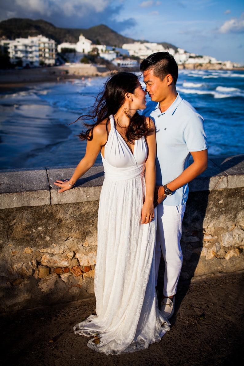 May include: A floor-length white dress with a halter neckline and a lace skirt paired with a light blue polo shirt and white trousers. The clothing is worn by two individuals standing together near a stone wall overlooking the ocean.