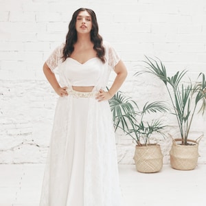 May include: A white lace bridal gown with a fitted bodice and a flowing skirt. The gown has a delicate lace overlay and a simple white belt.