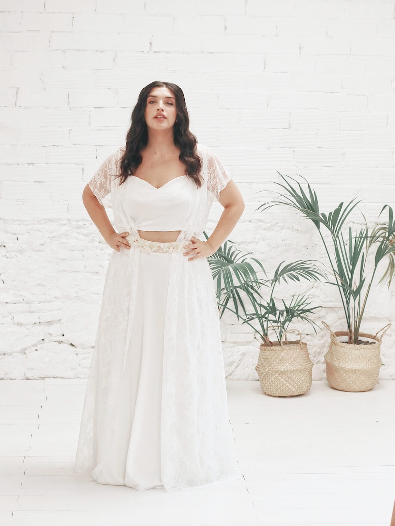 May include: A white lace bridal gown with a fitted bodice and a flowing skirt. The gown has a delicate lace overlay and a simple white belt.